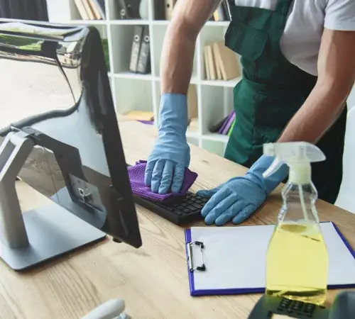 Cleaning technician wearing gloves disinfects keyboard and desk with spray bottle in bright office environment