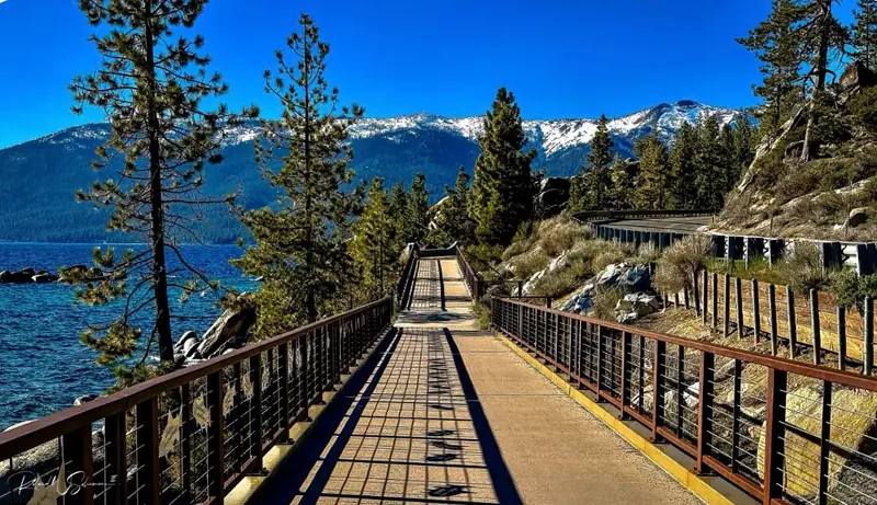 Incline Village lakefront boardwalk with railing, pine trees, and snow-capped mountains on a clear blue day