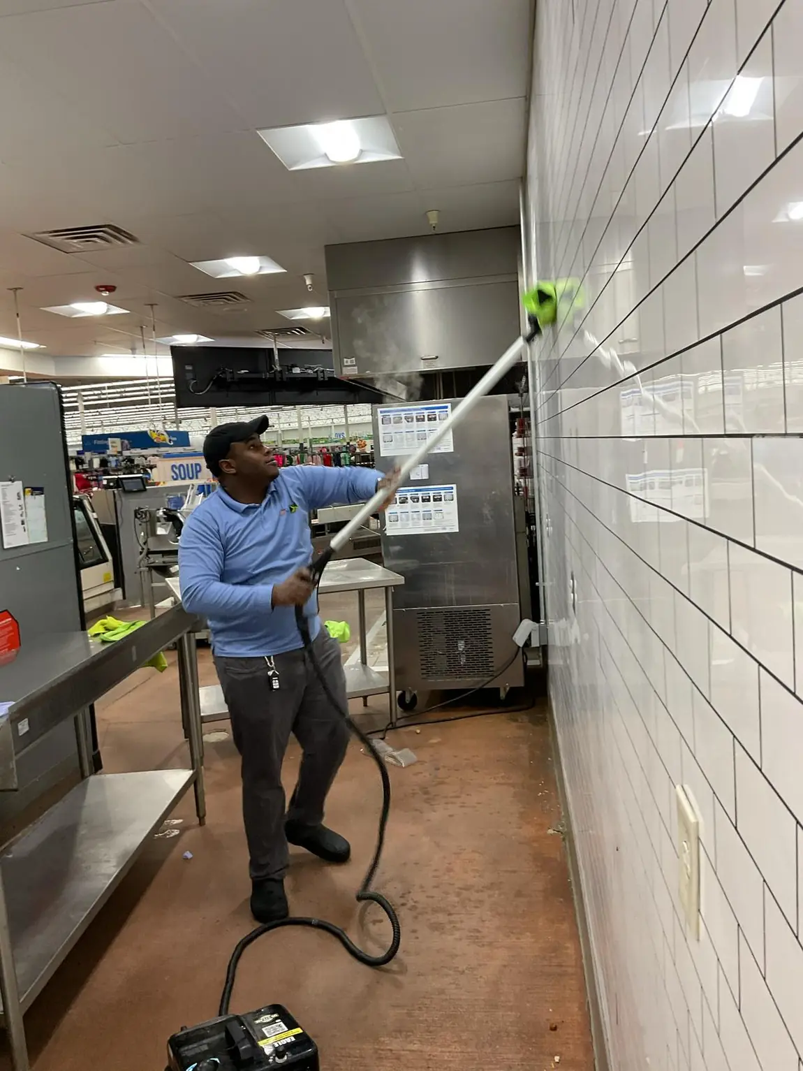 Janitorial cleaner scrubbing tiled kitchen wall with an extended pole scrubber in store kitchen