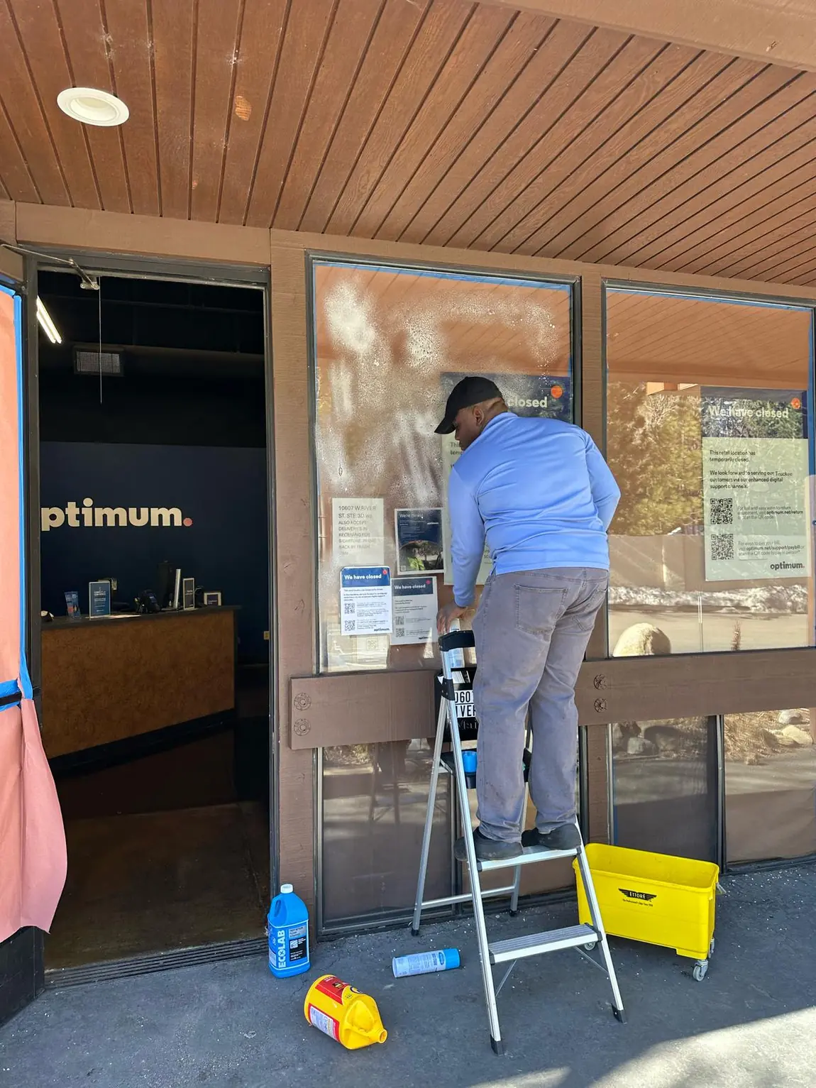 Technician performing commercial window cleaning on a storefront glass door and window using ladder and cleaning supplies