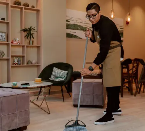 Commercial cleaning staff sweeping hardwood floor in a modern office lounge area with seating and shelving
